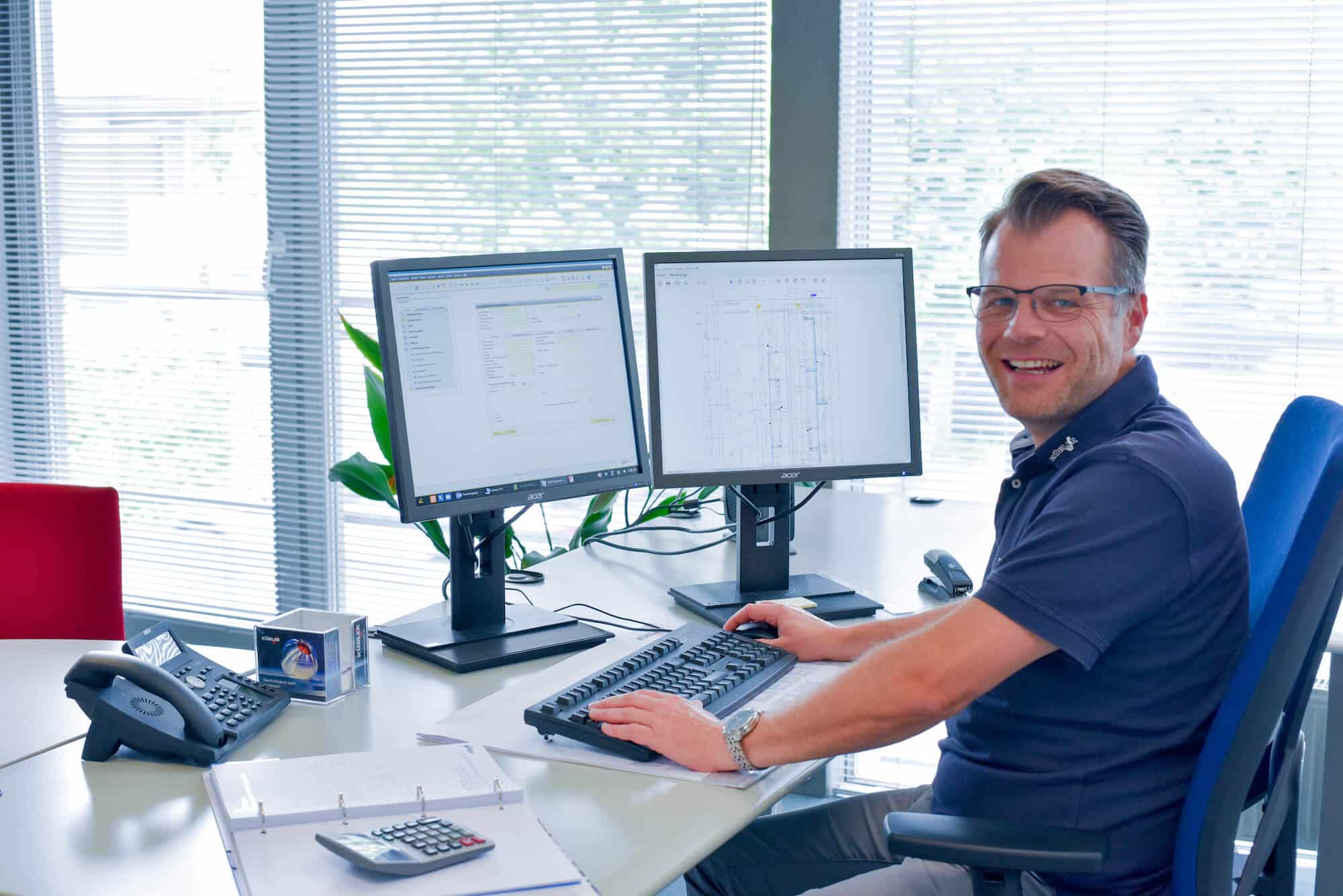 A KÜBLER employee sits laughing in front of two monitors and is working on a project.